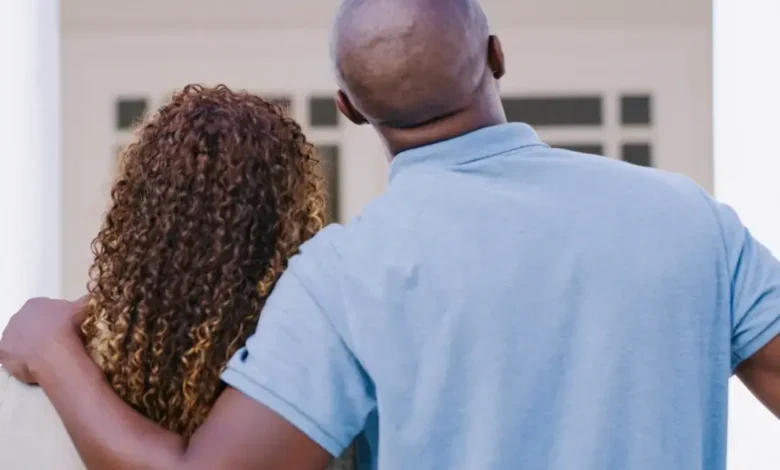 A couple stands with their backs to the camera, looking at a house, with the man's arm around the woman's shoulders, contemplating mortgage rates.
